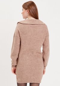 Robe en tricot beige avec un large col, des manches longues et une coupe cintrée, présentant une texture côtelée et une légère fente sur le côté.