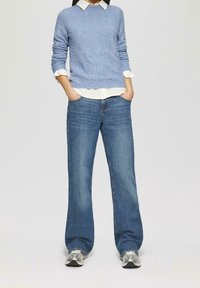 Maglione azzurro chiaro a trecce indossato sopra una camicia bianca con colletto, abbinato a jeans blu a gamba larga e sneakers argentate.