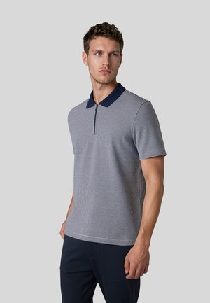 Mann, der ein kurzärmeliges Polohemd mit marineblauem Kragen und dünnen horizontalen Streifen trägt, steht vor einem einfarbigen hellgrauen Hintergrund.