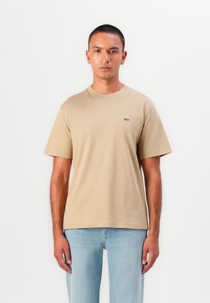 T-shirt en coton beige à manches courtes, col rond et un petit logo de crocodile vert sur la poitrine gauche. Porté avec un jean bleu clair.
