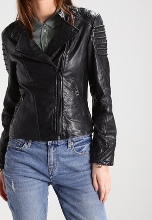 Frau trägt eine schwarze Lederjacke mit gepolsterten Schultern, Reißverschluss vorne und blaue Jeans vor einem einfarbigen hellen Hintergrund.
