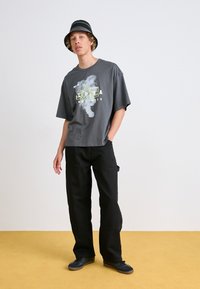 T-shirt graphique gris avec un design vert clair, associé à un pantalon cargo noir. Le mannequin porte un chapeau seau rayé noir et blanc et des baskets.