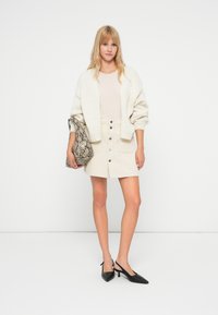 Femme portant un cardigan crème, une jupe beige boutonnée sur le devant, des talons slingback noirs, tenant une grande pochette à motif peau de serpent.