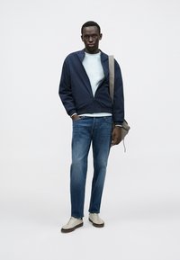 Marineblauwe bomberjas over een lichtblauw sweatshirt, gecombineerd met blauwe spijkerbroek en beige sneakers. Een bruine schoudertas maakt de look compleet.