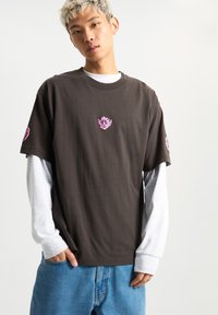 Camiseta marrón de manga corta con un diseño gráfico rosa en el pecho y la manga izquierda, usada sobre una camisa de manga larga gris, combinada con jeans azules.