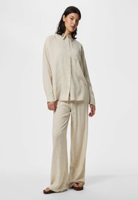 Femme debout portant une chemise beige ample boutonnée et un pantalon large assorti avec des sandales marron sur fond blanc.