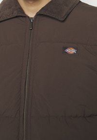 Brązowa kurtka puchowa z kołnierzem ze sztruksu, zamkiem błyskawicznym z przodu i haftowaną naszywką z logo Dickies po lewej stronie.