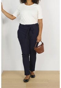 Mørkeblå bukser med en bindebælte, parret med en hvid kortærmet top. Holder en lille brun læderhåndtaske. Flade sorte sko.