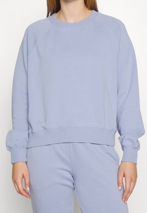 Woman wearing a light blue long-sleeve pullover sweatshirt and matching pants, standing with arms relaxed by sides.