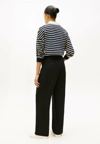 Frau mit einem Dutt, die einen navy- und weiß gestreiften, kurz geschnittenen Pullover, schwarze weite Hosen und weiße Sneaker trägt, steht mit dem Rücken zur Kamera.