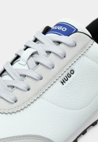 Baskets en cuir blanc et gris avec des lacets gris clair et une étiquette bleue sur la languette, avec la marque "HUGO" sur le côté.