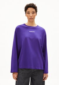 Lila langärmliges T-Shirt mit Rundhalsausschnitt, weichem Stoff und weißem "ARMEDANGELS"-Schriftzug auf der Brust. Getragen mit dunklen Jeans.