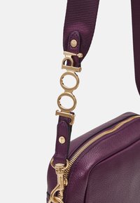 Sac en cuir violet avec une fermeture éclair dorée. La sangle ajustable présente un fermoir en métal doré et des détails de logo. La surface texturée ajoute de la profondeur.