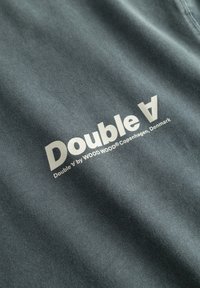 Szary materiał o gładkiej teksturze z białym logo: "Double V by WOOD WOOD Kopenhaga, Dania" wyraźnie wyeksponowane.
