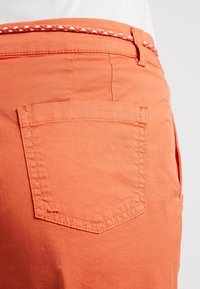Pantaloni corti in cotone arancione con tasca posteriore e una cintura intrecciata a contrasto. Texture liscia e design casual con dettagli di cucitura pulita.