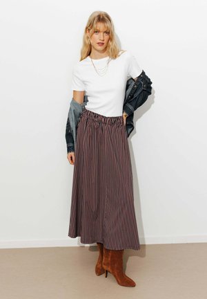 Femme blonde portant un haut blanc à manches courtes, une veste en jean noire, une jupe midi rayée, des bottines marron à talons et des colliers dorés superposés, debout à l'intérieur.