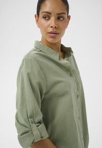 Femme portant une chemise vert clair à boutons, manches retroussées, regardant par-dessus son épaule sur fond blanc uni.