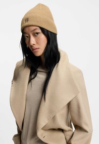 Ribattu beige neulepipo, jossa on logoyksityiskohta. Mallilla on päällään pehmeä, vaalea beige ylisuuri takki ja sen alla istuva beige paita.