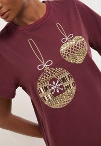 Burgundi T-särk kuldse ja hõbedase kaunistusprindiga, millel on kaks dekoratiivset palli, millel on detailne joonistus ja geomeetrilised mustrid.