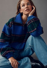 Fleece-Pullover mit Reißverschlusskragen, mit einem blau-grün-burgunderfarbenen Karomuster; kombiniert mit hellblauen Jeans und schwarzen Stiefeln.