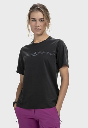 Frau mit zurückgebundenem hellem Haar, trägt ein schwarzes T-Shirt mit Grafik und lila Hose, steht mit den Händen in den Taschen vor einem schlichten Hintergrund.