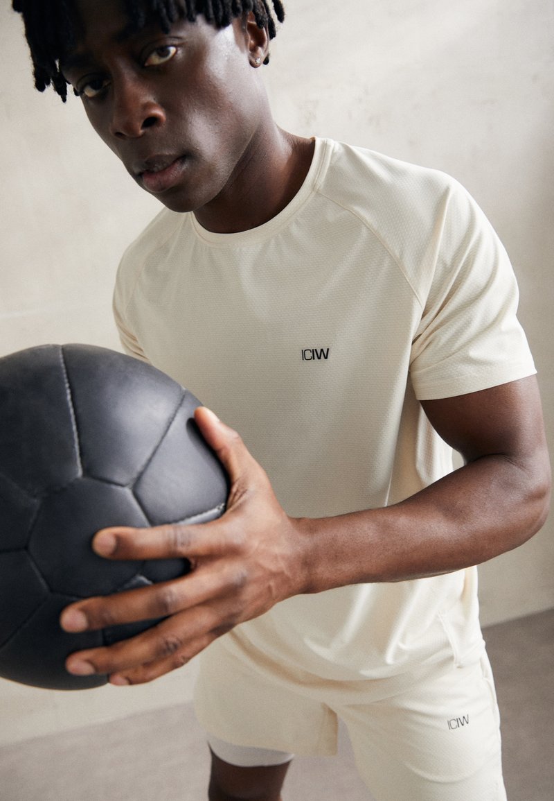 Camisa atlética de color beige claro con tejido texturizado y costuras en los hombros, combinada con pantalones cortos a juego. El modelo sostiene un balón de fútbol negro.