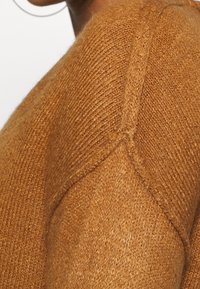 Pull marron tricoté avec une finition texturée, présentant un détail de couture le long de l'épaule. Le matériau semble doux et chaud.