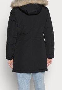 Svart huva parka med fuskpälskant, med ett vadderat design och långa ärmar. Silhuetten är avslappnad, med en midjehöjd längd.