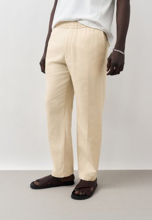 Άτομο που φοράει μπεζ λινά παντελόνια με ελαστική μέση, λευκό πουκάμισο, ασημένιο βραχιόλι και σκούρα καφέ σανδάλια με ανοιχτά δάχτυλα σε γκρι ανοιχτό πάτωμα.