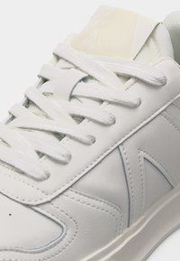 Detailný záber bielej tenisky so zaviazanými šnúrkami, zobrazujúci precízne prešívanie, hladký kožený materiál a polstrovaný jazyk.