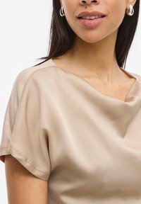Blouse en satin beige à manches courtes, avec un décolleté drapé et une texture lisse. Présente des détails de couture subtils le long des bords.