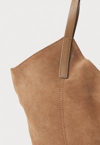 Bolso tote de ante en marrón claro con un acabado texturizado. Presenta un asa única de cuero y costuras visibles a lo largo de los bordes.