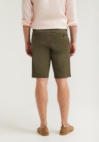 Olivgröna shorts med rak skärning, bakfickor med dragkedjor och en slät yta, tillsammans med bruna slip-on skor.