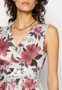 Kvinna som bär en ärmlös blommig klänning med stora rosa och vita blommor och ett tunt guldhalsband, visad från axlarna till midjan.