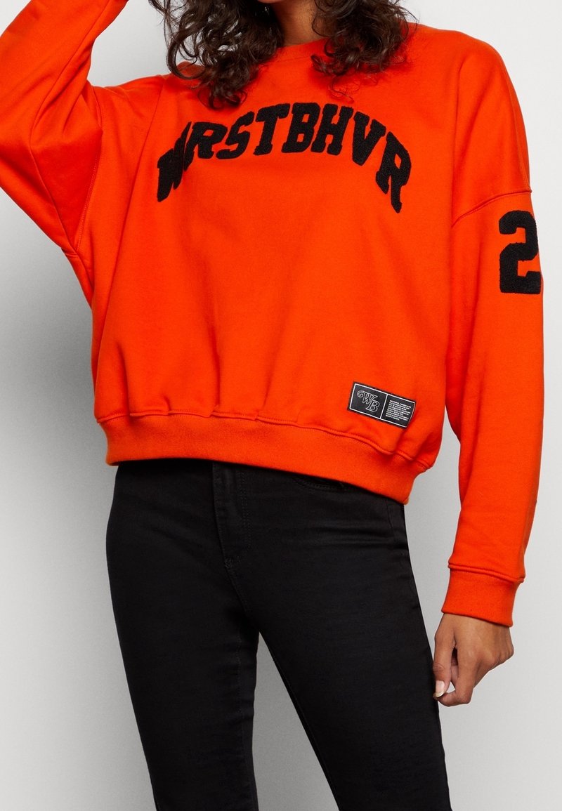 Personne portant un sweat-shirt orange vif avec le texte noir "WRSTBHVR" sur la poitrine, le numéro "2" sur la manche, et un pantalon noir.