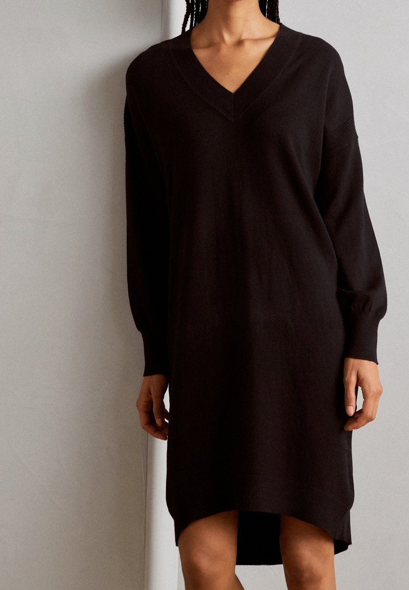 Abito nero lavorato a maglia con scollatura a V, maniche lunghe e polsini a costine; presenta un orlo leggermente curvato e una vestibilità comoda.