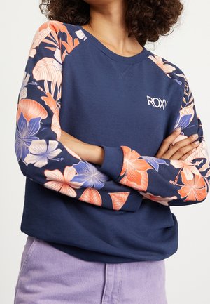 Kobieta nosząca granatową bluzę z pomarańczowymi i fioletowymi kwiatowymi rękawami oraz logo „ROXY”, w komplecie z jasnofioletowymi spodniami.
