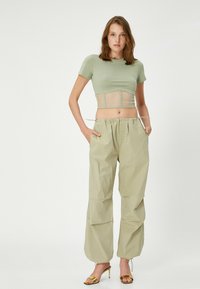 Lys grøn cropped t-shirt med en gennemsigtig mesh-detalje ved taljen, parret med løse, lyse beige bukser med lommer. Gyldne strophæle.