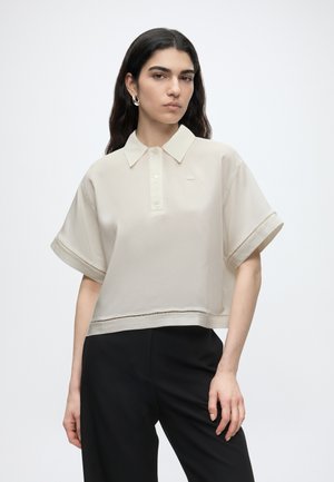 Femme aux longs cheveux foncés portant une chemise beige à manches courtes avec col et un pantalon noir, debout devant un fond clair uni.