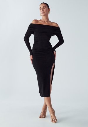 Zwarte off-the-shoulder jurk met lange mouwen, geplisseerd ontwerp en hoge split tot de dij. Draag met gouden sandalen met banden. Gladde stoffen textuur.