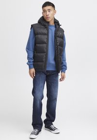 Schwarze gefütterte Weste mit Kapuze über einem blauen Sweatshirt, kombiniert mit blauen Jeans und schwarzen Sneakers. Mit gestepptem Design und Reißverschlussdetails.