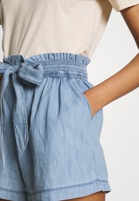 Ljust blå chambrayshorts med elastisk midja, knytdetalj och sidfickor. Slät textur, avslappnad design och fransad nederkant.