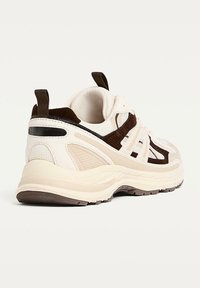 Chaussure de sport beige avec des accents marron, en maille texturée et matériaux synthétiques, dotée d'une semelle épaisse et d'une languette à l'arrière pour faciliter l'enfilage.