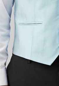 Világoskék, textúrált mellény, finom gyémántmintával, fehér inggel párosítva. Egyetlen zsebe van, és sötét nadrág felett viselik.
