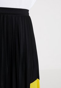 Gonna nera plissettata con una fascia larga in vita e un pannello colorato giallo nella parte inferiore. Tessuto leggero e strutturato.