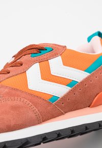 Baskets orange et marron avec une tige en mesh et suède, un motif zigzag blanc, des accents bleu sarcelle et une semelle en caoutchouc noire.