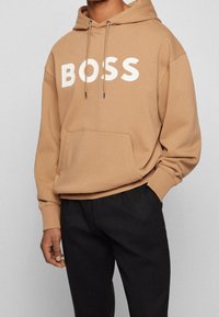 Persona vestida con sudadera beige con el texto blanco "BOSS" y pantalones negros, de pie con una mano en el bolsillo frente a un fondo liso.