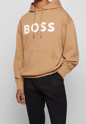 Persoon in een beige hoodie met witte "BOSS" tekst en zwarte broek, staat met één hand in de zak tegen een effen achtergrond.