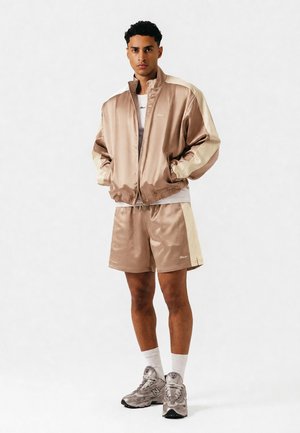 Mann står med hendene i lomma, iført beige satin treningsjakke og matchende shorts, hvite sokker og grå joggesko mot hvit bakgrunn.