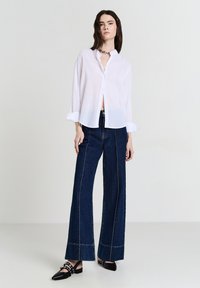 Camicia bianca con bottoni e maniche lunghe, abbinata a jeans larghi blu scuro con cuciture a contrasto e scarpe nere con punta.